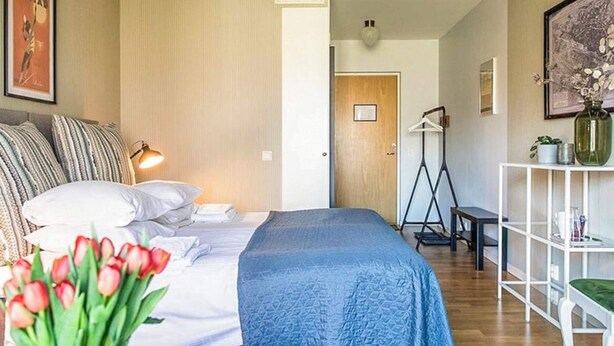 A bedroom with a bed, pillows, a bedside lamp, a desk, a chair, a shelf with decorative items, and a door leading to another room. - 2 Person Holiday Park Home in Bastad (Bastad)