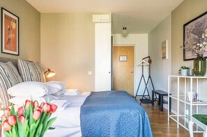 Un dormitorio con cama, almohadas, lámpara de noche, escritorio, silla, estante con adornos y puerta que da a otra habitación.
