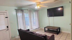 A room with a ceiling fan, a flat-screen TV mounted on the wall, a window with curtains, a door, and a small table. - Spacious 3-bedroom house in brilliant Austin  /The Domain & Q2 Stadium/ (Austin)