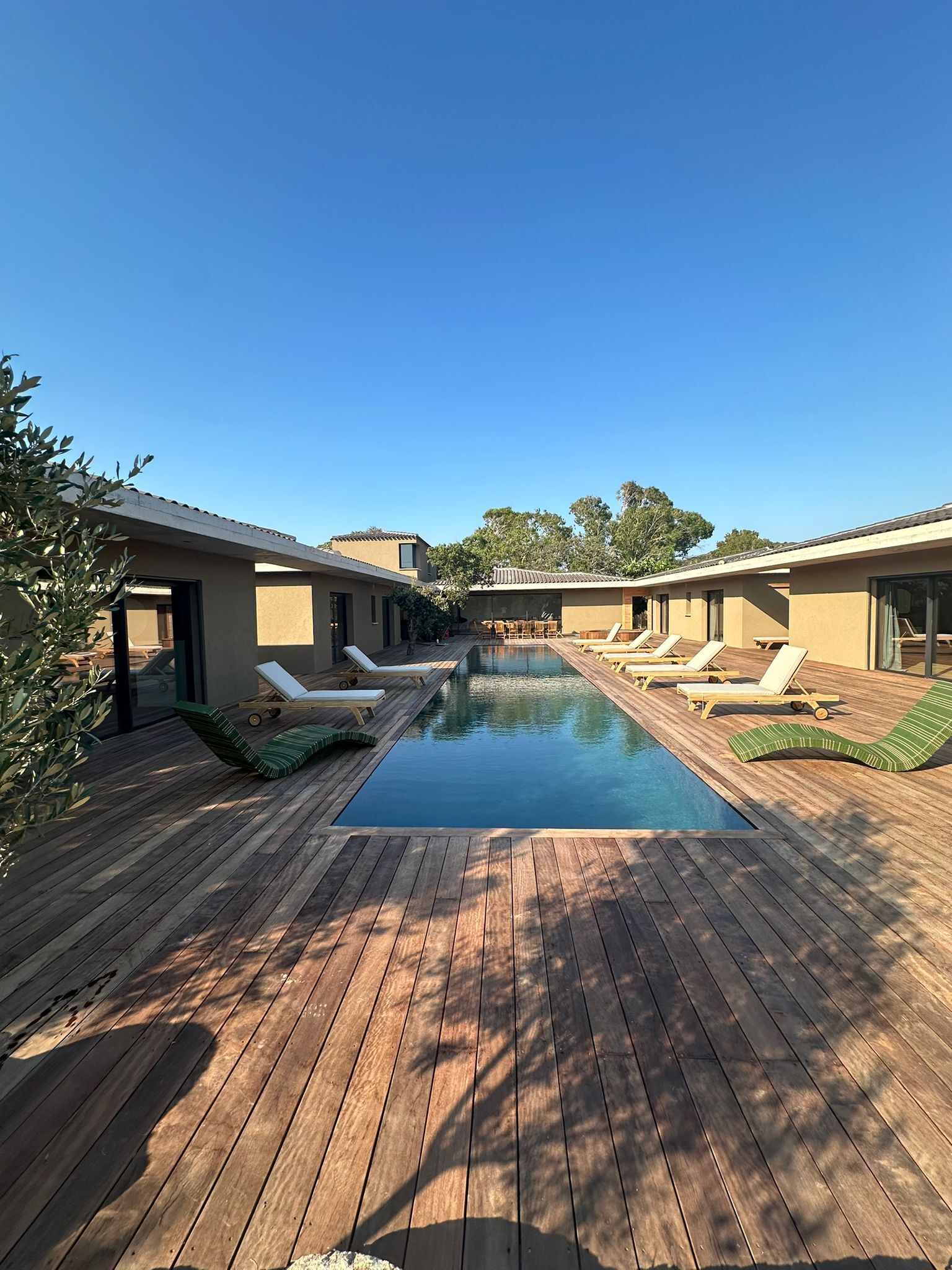 Un área de piscina con cubierta de madera, sillones de descanso y cielo azul despejado.