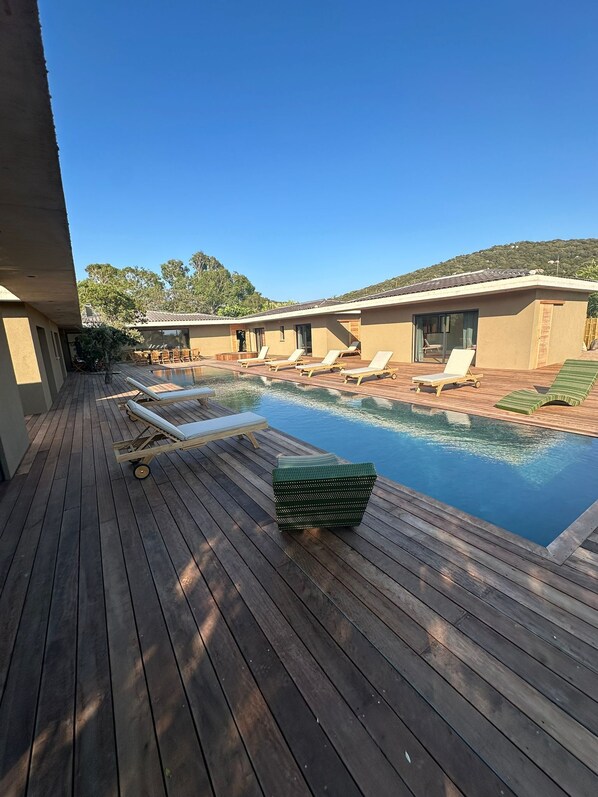 A wooden deck with a pool, lounge chairs, and a small building with a tiled roof.