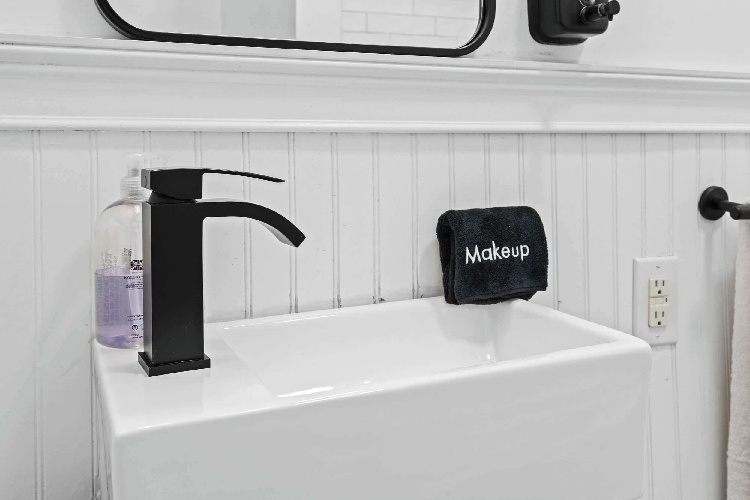 Una bañera blanca con un grifo negro, un perchero negro con una toalla que dice 'Maquillaje', y una botella de jabón líquido.