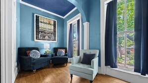 Ein Zimmer mit blauer Decke, einem großen Fenster mit Vorhängen, einem blauen Sessel, einer Couch und einem im Wand eingelassenen farbigen Glasfenster.