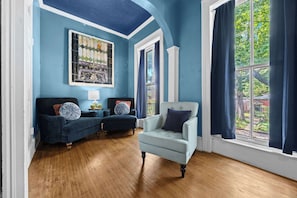 A room with a blue ceiling, a large window with curtains, a blue armchair, a sofa, and a wall-mounted stained glass window.