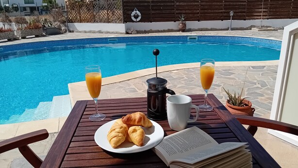 Une table au bord de la piscine, avec une assiette de croissants, un livre, une machine à café et deux verres de jus d’orange.