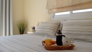 Un lit bien fait, avec une literie à rayures, une machine à café, une tasse de café et un panier contenant des croissants et un verre de jus d’orange, le tout posé sur une table.