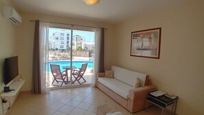 Un salon comprenant un canapé, un téléviseur, une petite table et un balcon avec vue sur la piscine.