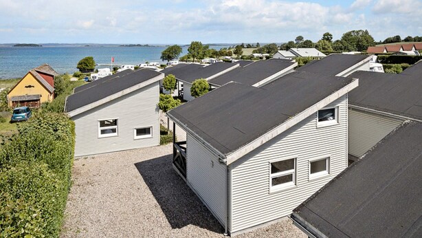 A row of modern, single-story mobile homes with a gravel driveway, set against a backdrop of a clear sky and a body of water. - 6 Person Holiday Park Home in Faaborg (Faaborg)