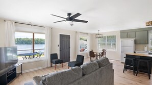 Amplia sala de estar con un sofá grande de color gris, un televisor y un ventilador de techo. Hay un comedor con una mesa y sillas, y una cocina con un refrigerador y armarios.