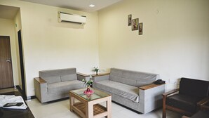 Een woonkamer met twee grijze banken, een glazen salontafel, een houten bijzettafeltje, een zwarte leren fauteuil en een airconditioner aan de muur.