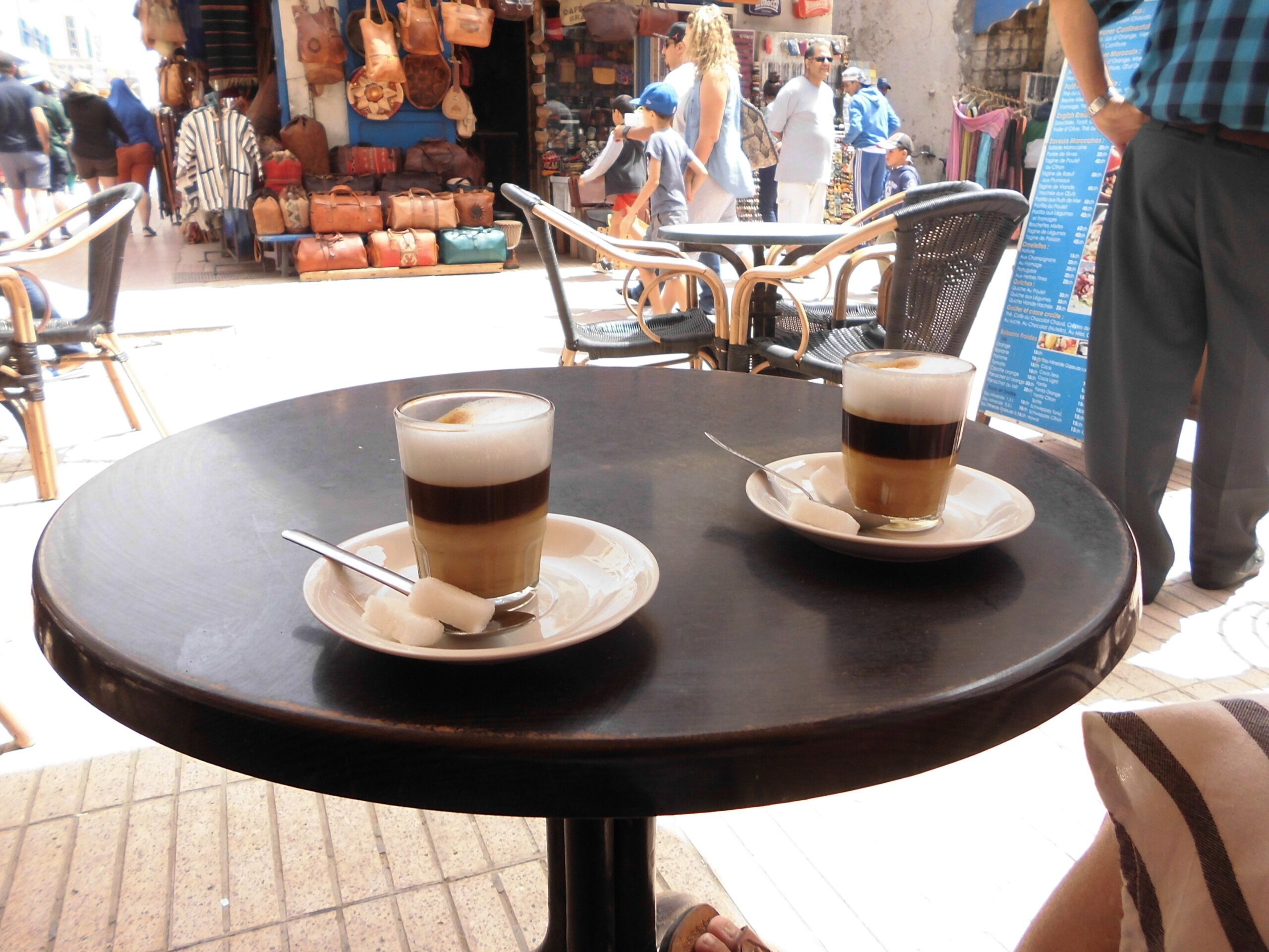 Two cups of coffee on a round table with a view of a busy street and shops in the background.