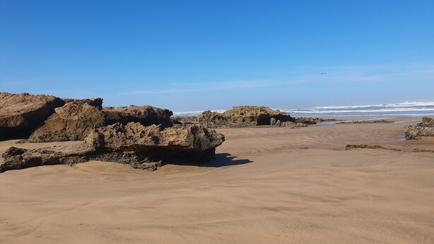 Une plage de sable fin, parsemée de grandes formations rocheuses, sous un ciel bleu et dégagé.