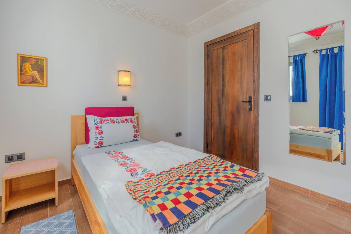 A bedroom with a bed, a wooden door, a wall-mounted light, a small bedside table, and a mirror reflecting a bathroom with a blue shower curtain.