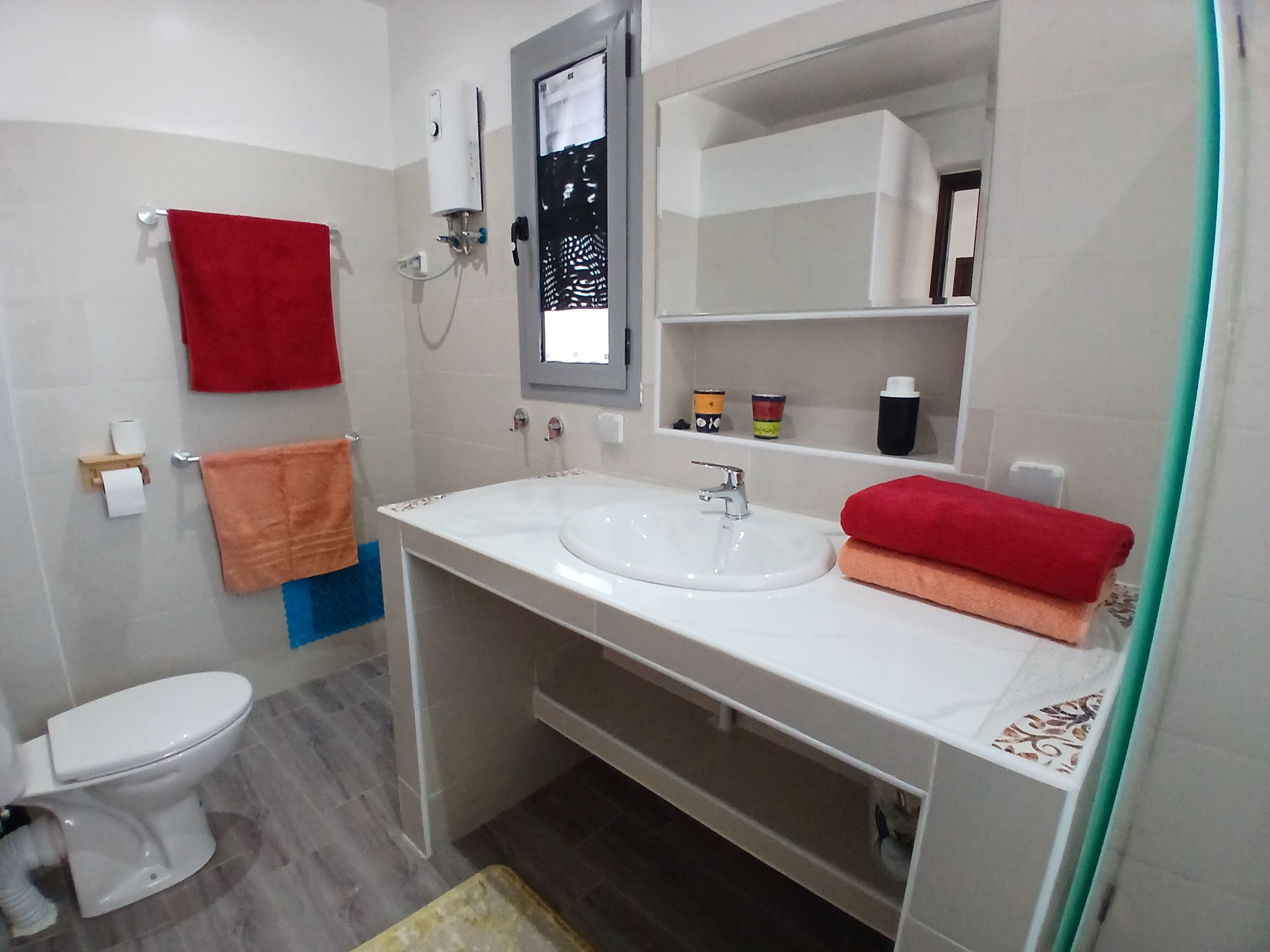 A bathroom with a white sink, a toilet, and a mirror above the sink. There are towels hanging on the wall and a shelf with toiletries.