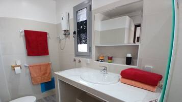 A bathroom with a white sink, a toilet, and a mirror above the sink. There are towels hanging on the wall and a shelf with toiletries.