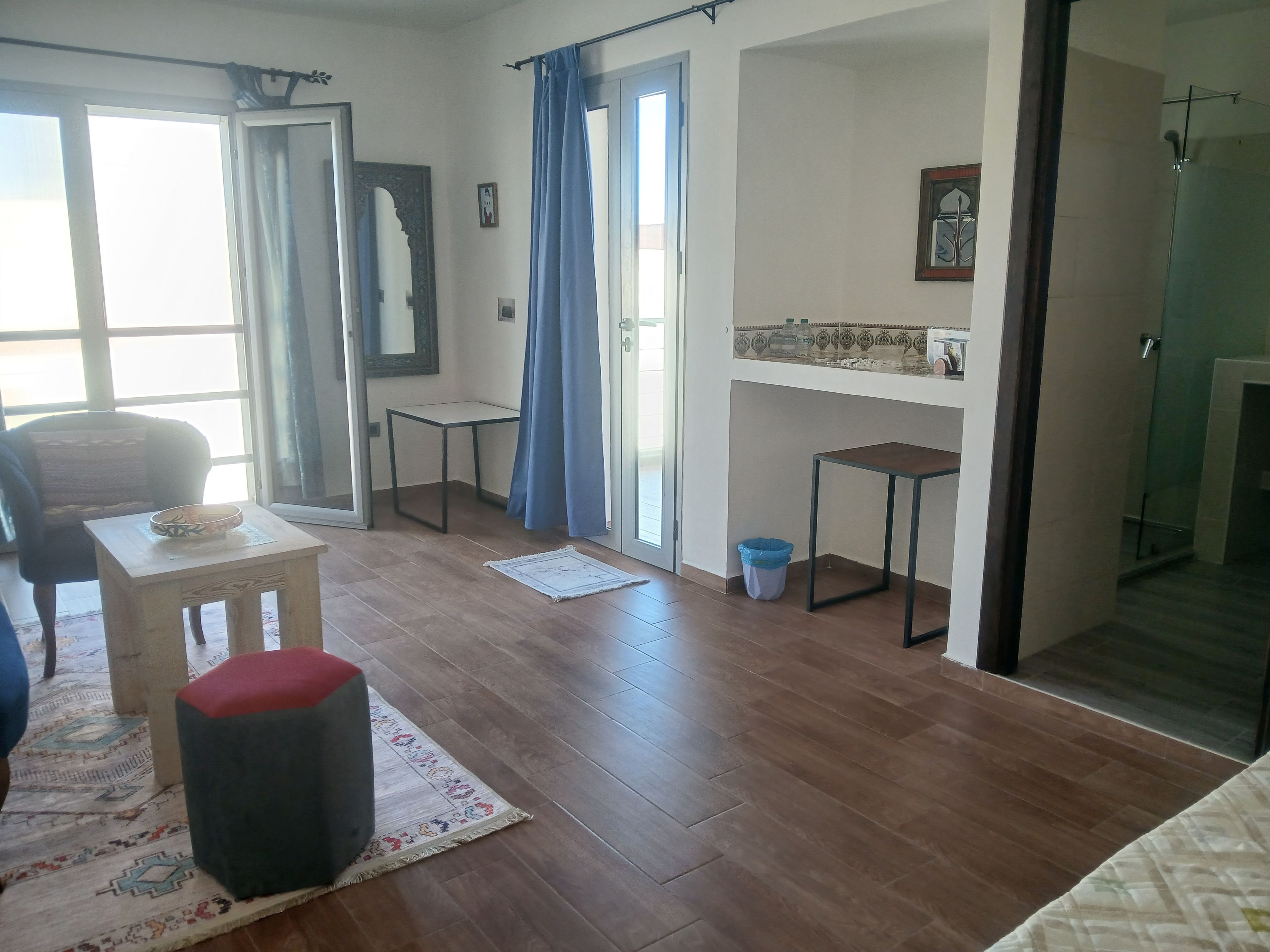 A room with a wooden floor, a small table with a bowl, a chair, a large mirror, a blue curtain, a small trash bin, and a glass shower area.