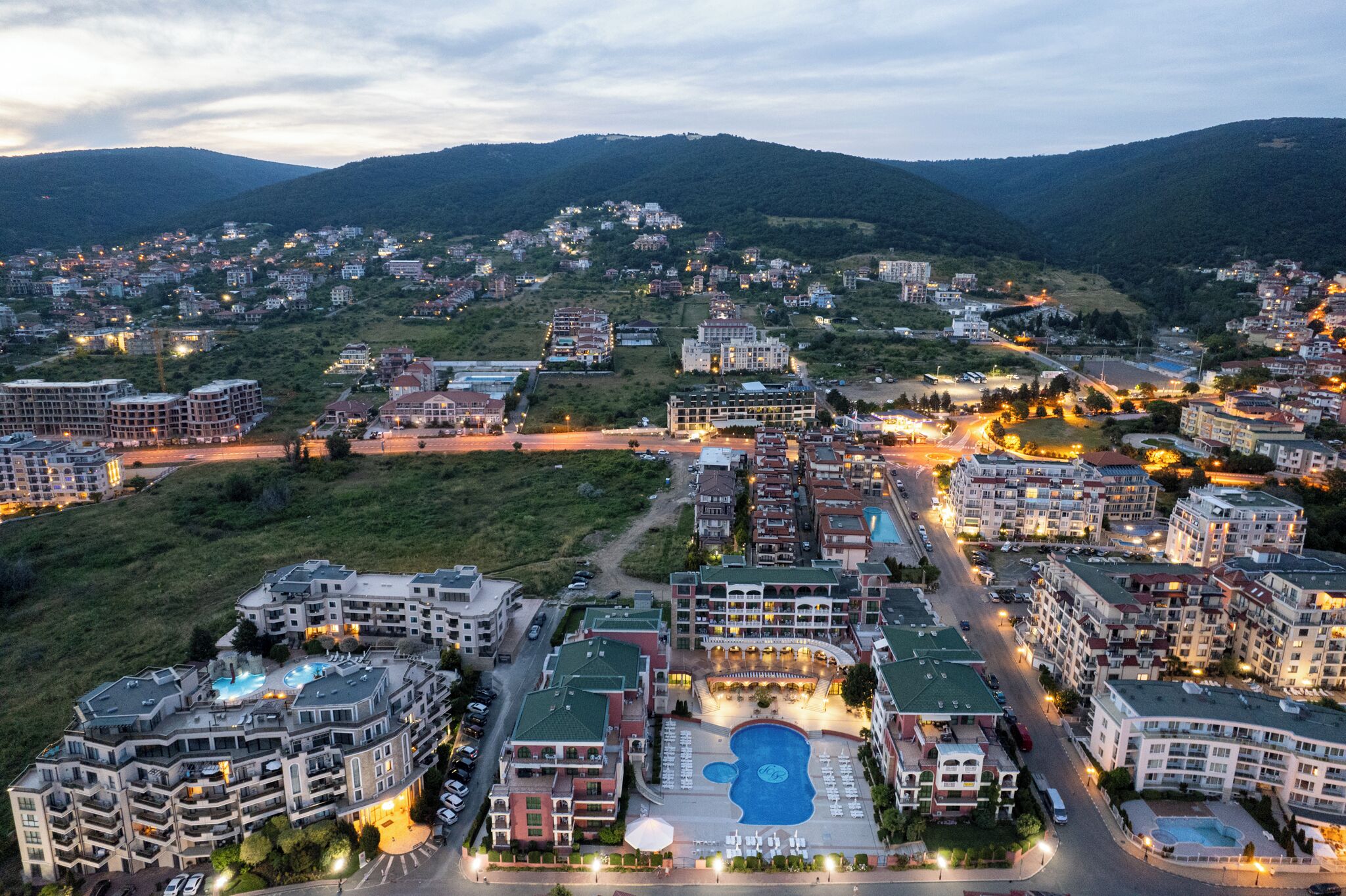 A residential area with multiple apartment buildings, a swimming pool, and a hilly landscape in the background.