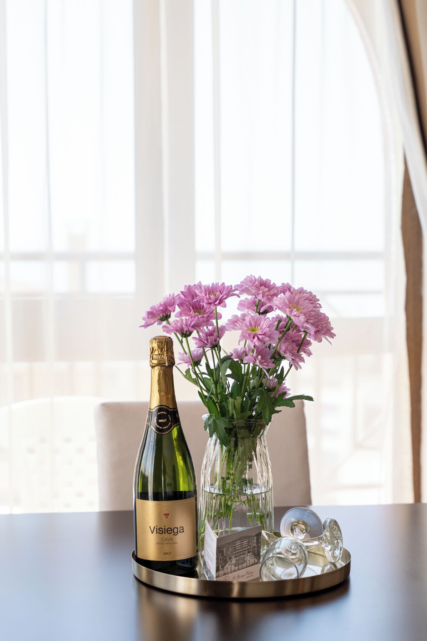 A bottle of Visiege Brut champagne, a glass vase with pink flowers, and a tray with glasses on a table by a window.