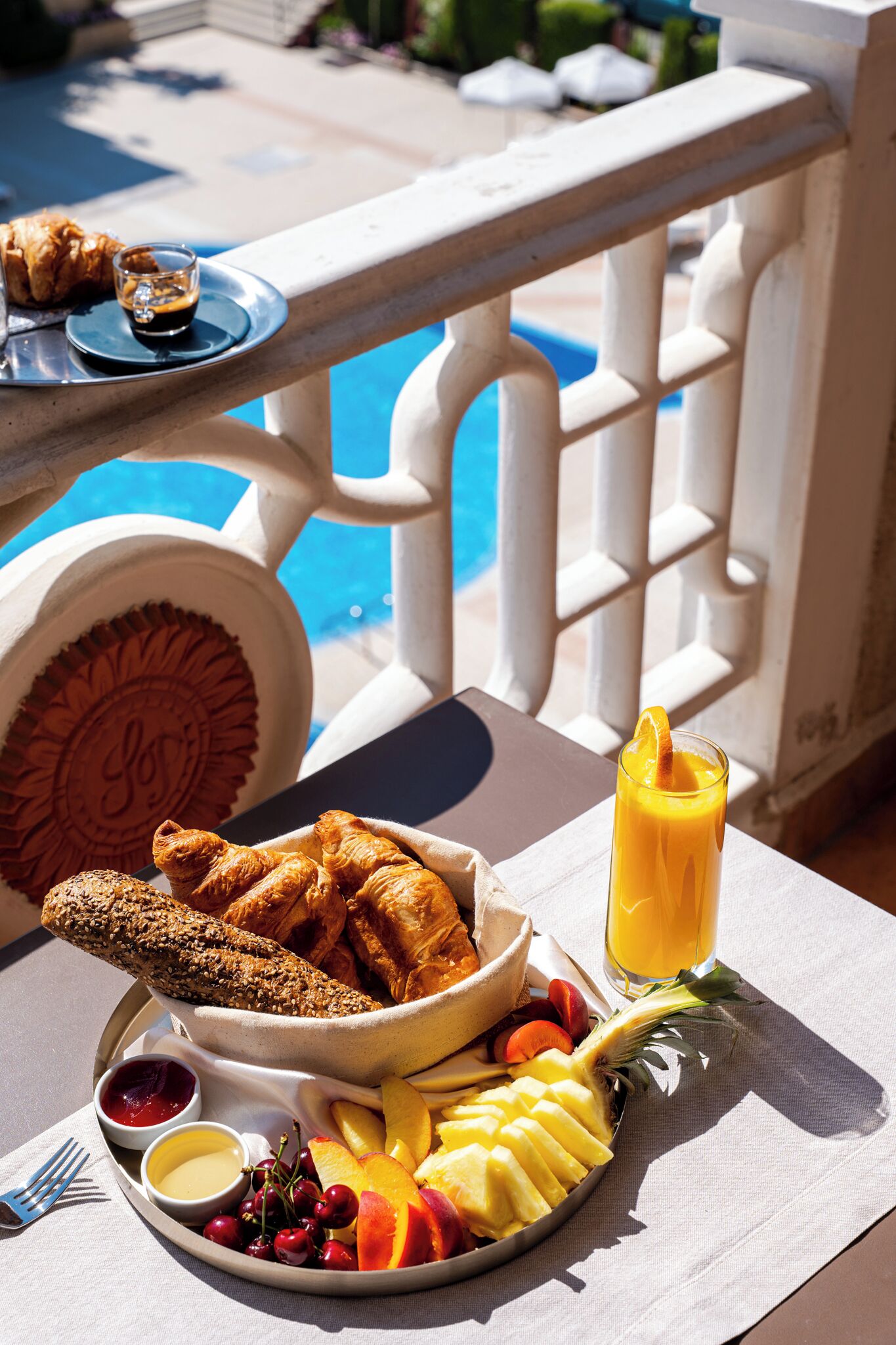 A dining table with a variety of food items including bread, fruits, and a glass of orange juice, set against a poolside view.