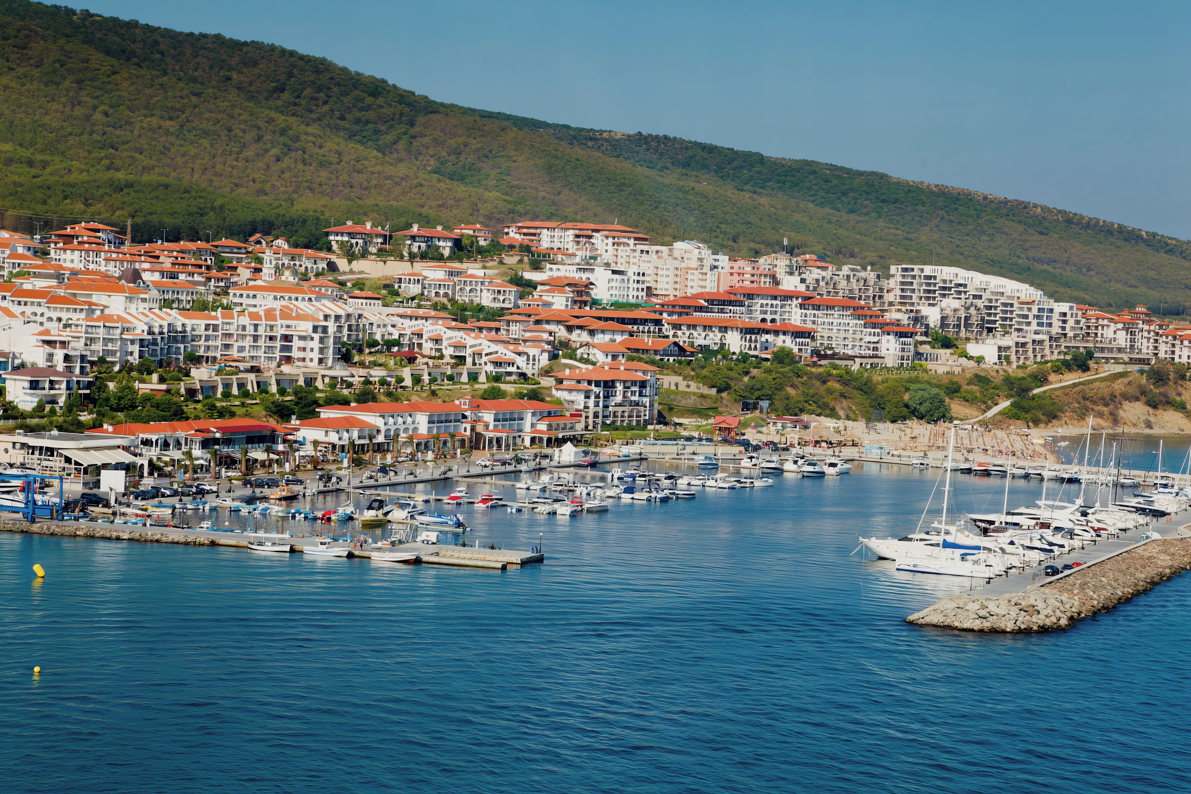 A coastal town with a marina, featuring multi-story buildings with red roofs, situated on a hillside.