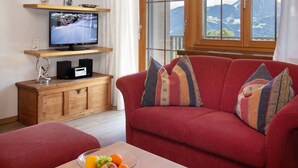 Ein Wohnzimmer mit einem roten Sofa, einem Couchtisch aus Holz mit einer Obstschale, einem Fernseher auf einem Holzständer und einem Fenster mit Blick auf die Berge.