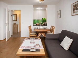 Un salon chaleureux avec un canapé gris foncé, une table à manger en bois mise avec des tasses et des soucoupes, et une cuisine visible à travers une porte ouverte.