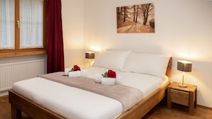 A neatly made bed with a wooden frame, two bedside tables with lamps, a window with sheer curtains, and a painting of a forest on the wall. - Nice apartment for 5 people with WIFI, pool, TV, balcony and panoramic view (Falera)