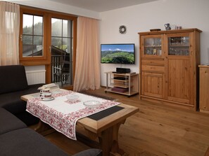 Un salon chaleureux avec un canapé, une table basse recouverte d’une nappe à motifs, un téléviseur, un meuble en bois et une fenêtre avec des rideaux.