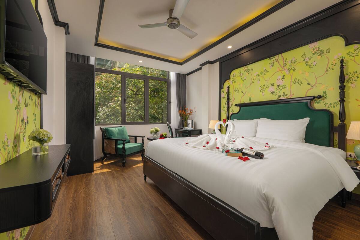 A bedroom with a large bed, a green chair, a desk, and a window with a view of trees.