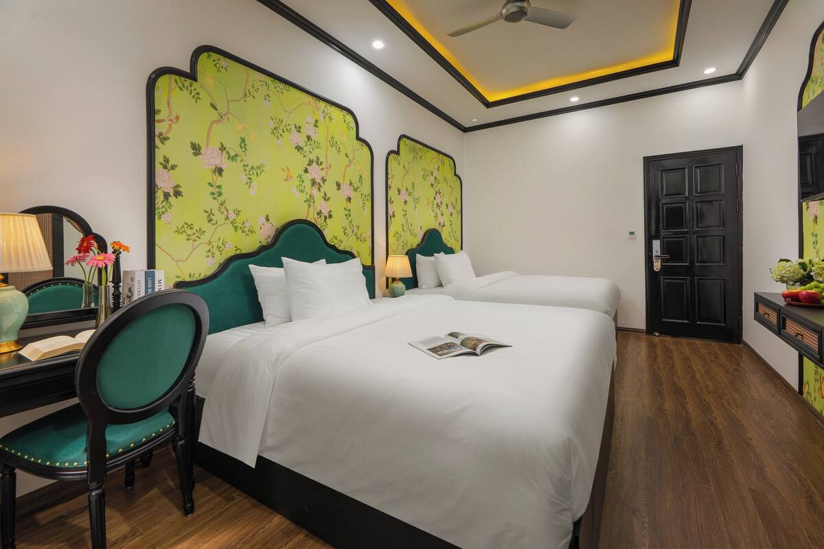 A hotel room with two beds, a desk with a chair, a desk lamp, and a bookshelf.