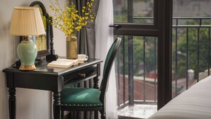 Una habitación con escritorio, lámpara, un jarrón con flores amarillas, una silla y vistas al exterior a través de una ventana.
