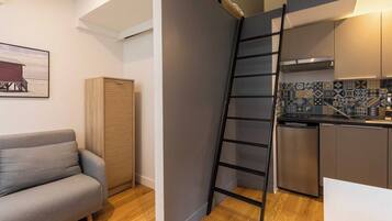 A compact living space with a grey sofa, a wooden cabinet, a kitchen with a tiled backsplash, and a black ladder.