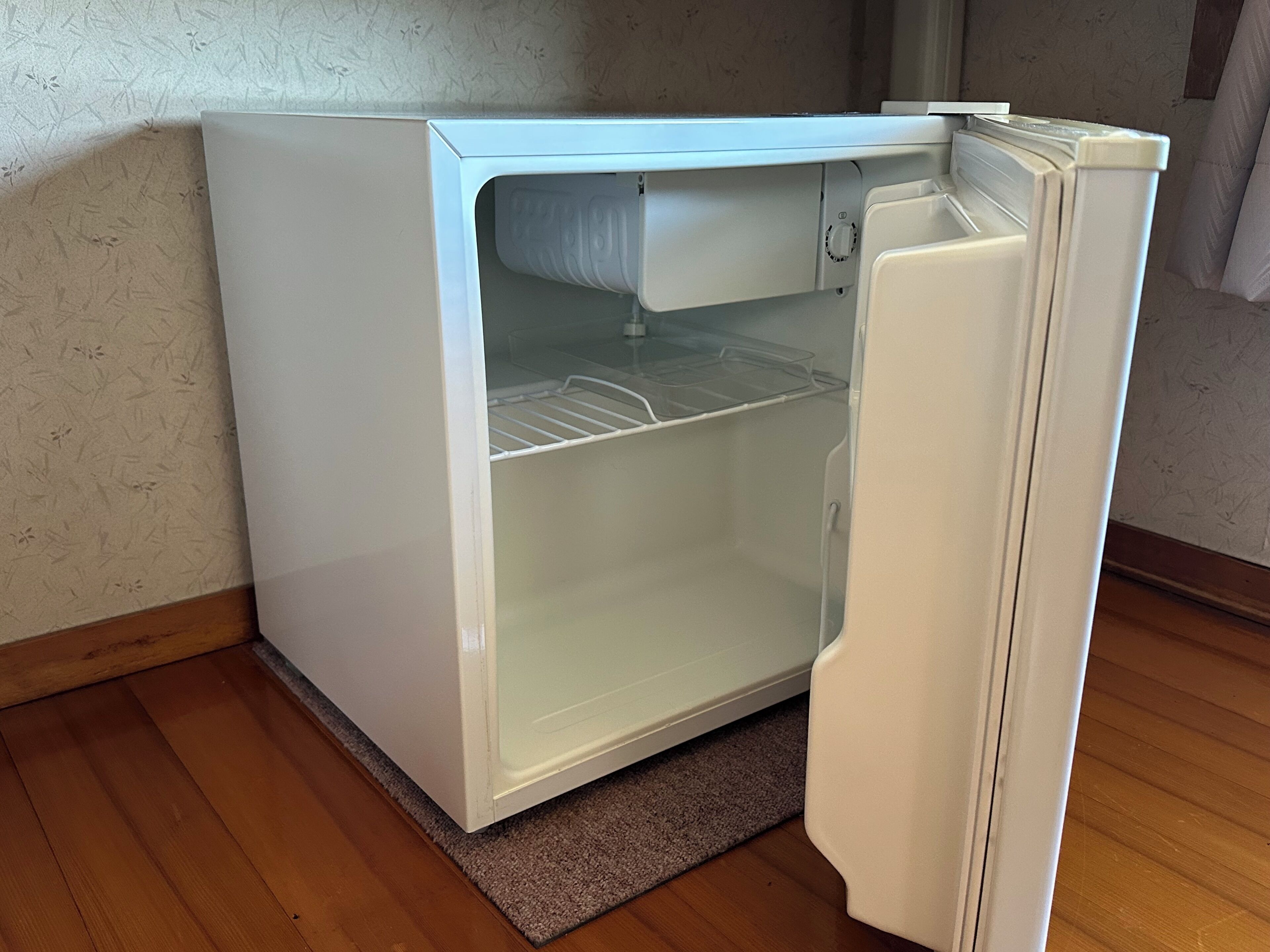 Un refrigerador blanco compacto con compartimento congelador, sobre un piso de madera frente a una pared texturizada.