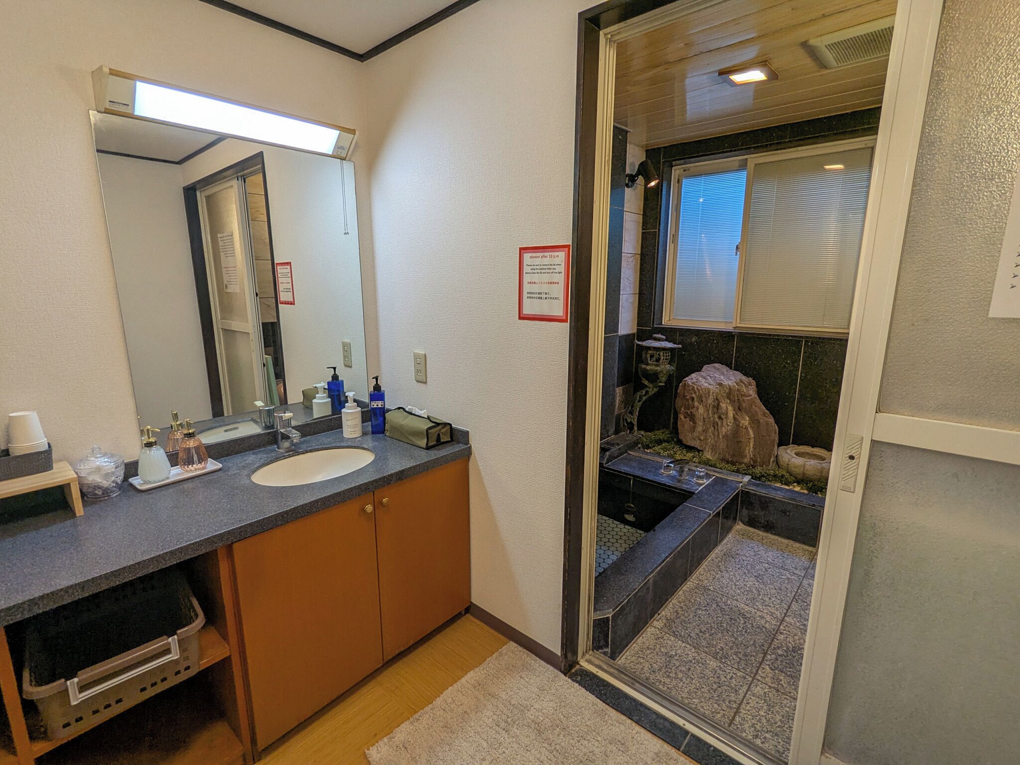 Een badkamer met een wastafel, spiegel en toiletartikelen. Er is een klein raam met zonwering en een decoratief rotsfragment.