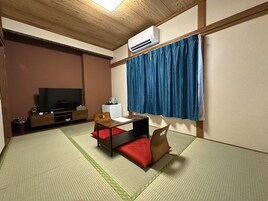 Una habitación de estilo japonés tradicional con suelo de tatami, una mesa baja con cojín, televisión y un refrigerador pequeño.