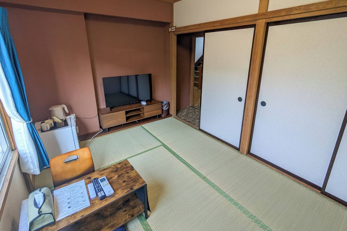 Una habitación tradicional japonesa con suelo de tatami, un televisor sobre un soporte de madera, una pequeña mesa con un control remoto y puertas correderas.