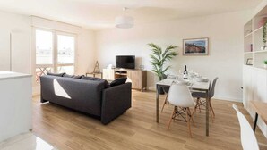 A modern living room with a sofa, dining table, and chairs. There is a TV, a plant, and a window with a view. - Dify Bollier - Quartier Gerland (Lyon)