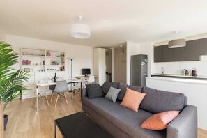 A modern living room with a grey sofa, a dining table, and a kitchen area with white cabinets and a grey refrigerator. - Dify Bollier - Quartier Gerland (Lyon)