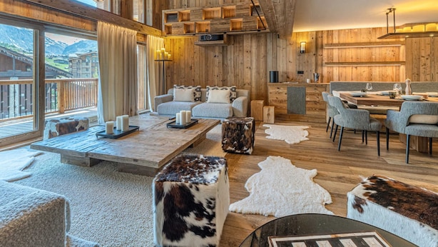 Ein großzügiger Raum mit hoher Decke, Holzbalken, einer gemütlichen Sitzecke mit Sofa und Sesseln, einem Esstisch für zwei Personen und Bergblick durch große Fenster.