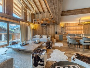 Ein großzügiger Raum mit hoher Decke, Holzbalken, einer gemütlichen Sitzecke mit Sofa und Sesseln, einem Esstisch für zwei Personen und Bergblick durch große Fenster.