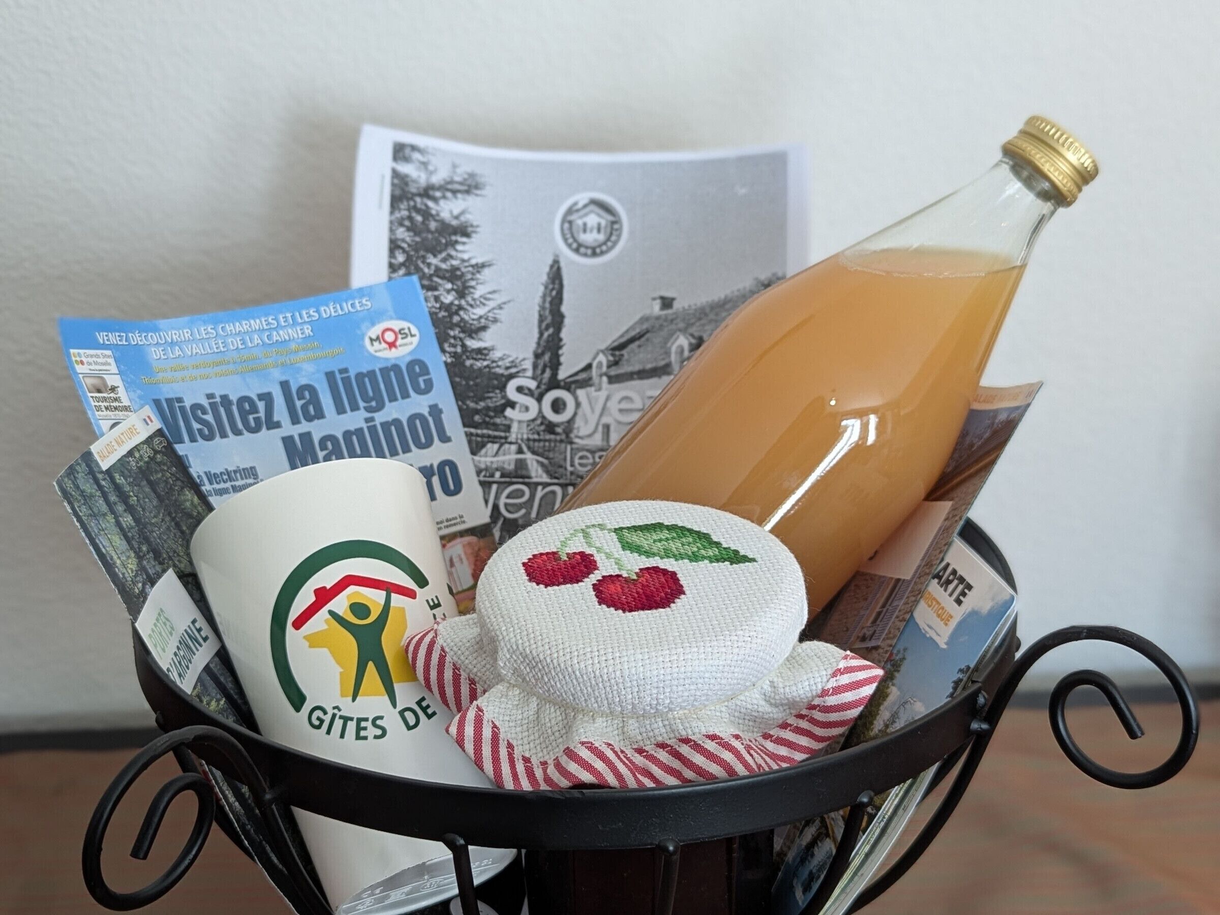Uma cesta com uma garrafa de suco de laranja, uma caneca branca com um logotipo verde, um tecido listrado em vermelho e branco e uma revista com capa francesa.