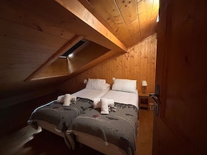 Um quarto de sótão aconchegante, com teto inclinado, paredes de madeira, uma cama de casal com duas almofadas, um abajur e uma pequena mesa.