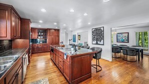 A modern kitchen with wooden cabinets, a central island, and a dining area with a view of trees. - Black Bear Ridge | Games, Hot Tub, Deck w/ Views (Mentone)