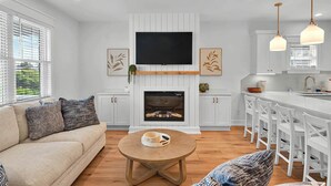Een moderne woonkamer met een flatscreen televisie, een open haard, witte kasten en een eethoek met barstoelen.