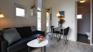 Un salon moderne avec un canapé noir, une petite table ronde sur laquelle se trouve un saladier rempli de fruits, un coin repas avec une table et des chaises, et une cuisine visible à travers une porte ouverte.