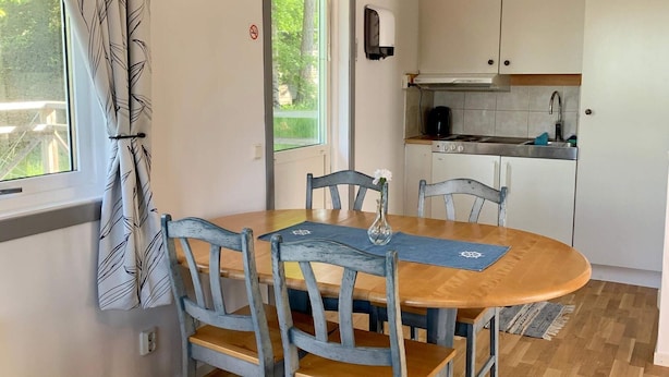 A dining area with a wooden table and chairs, a vase with flowers, and a view of the kitchen in the background.