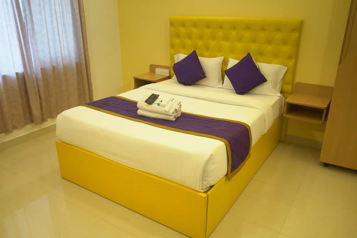 Habitación de hotel con una cama amarilla, ropa de cama blanca, almohadas moradas, un control remoto y una toalla doblada sobre la cama.
