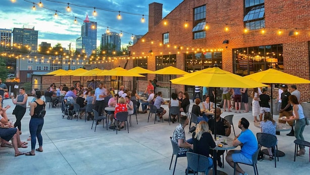 An outdoor patio area with tables and chairs, yellow umbrellas, and string lights against a brick building backdrop. - Across Raleigh Convention Center + Dining & Pool (Raleigh)