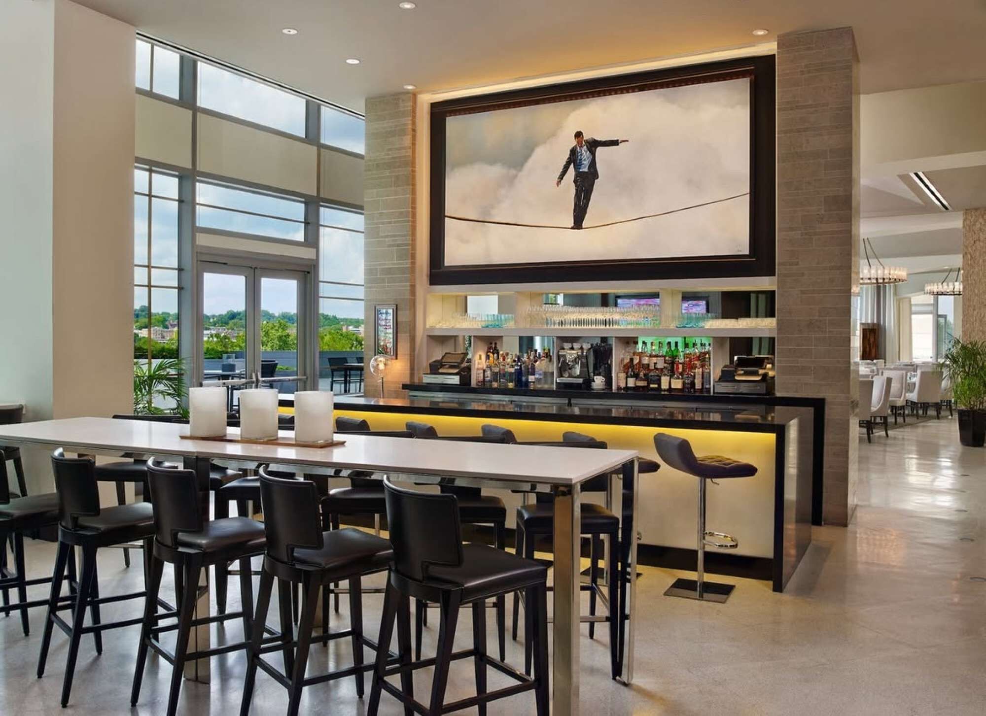 Zona de bar moderna con una pantalla grande que muestra a un hombre caminando sobre una cuerda alta, taburetes de bar y vistas al exterior a través de amplias ventanas.