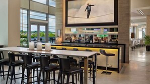 Zona de bar moderna con una pantalla grande que muestra a un hombre caminando sobre una cuerda alta, taburetes de bar y vistas al exterior a través de amplias ventanas.
